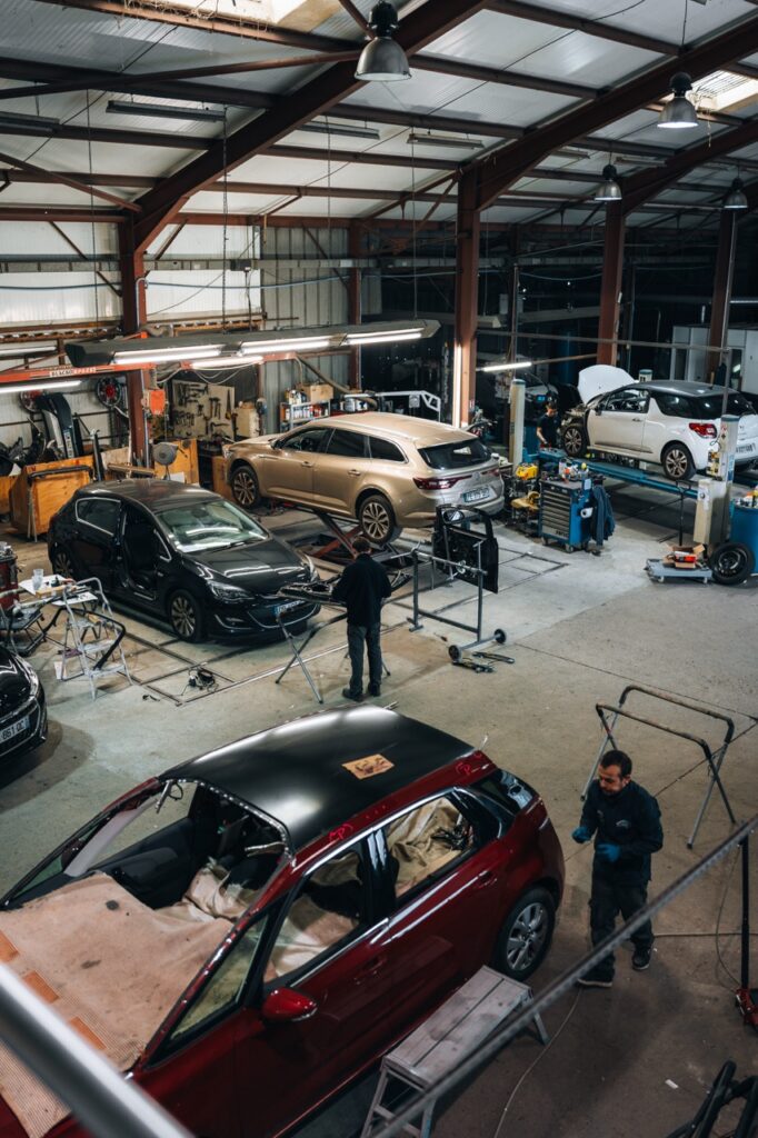 Atelier de réparation automobile animé avec plusieurs mécaniciens travaillant sur des voitures, dont une station wagon sur pont et une voiture rouge au toit noir décapée.