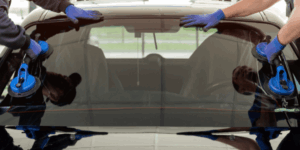 Deux techniciens en gants bleus installent le pare-brise arrière d'une voiture noire avec des ventouses. Remplacement vitre auto.