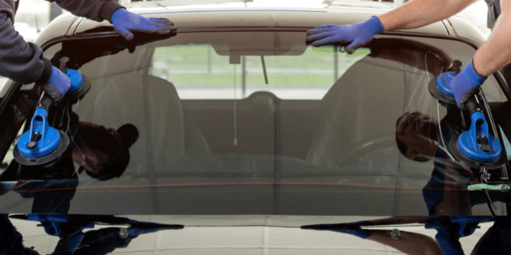 Deux techniciens en gants bleus installent le pare-brise arrière d'une voiture noire avec des ventouses. Remplacement vitre auto.